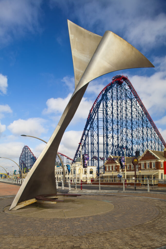 England, Lancashire, Blackpool. Whale tail sculpture on Blackpool's South Promenade with the Blackpool Pleasure Beach and the Big One (steel roller coaster) in the distance.