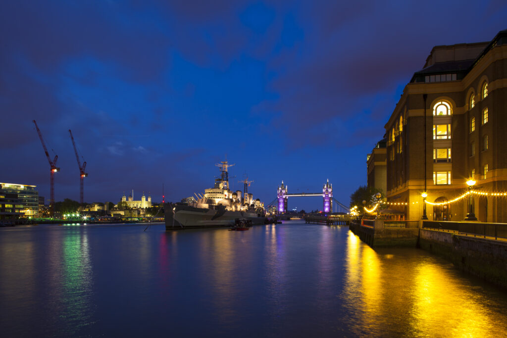 England, London, Southwark. HMS Belfast, originally a Royal Navy light cruiser, permanently moored in London on the River Thames with Tower Bidge and the Tower of London in the distance.