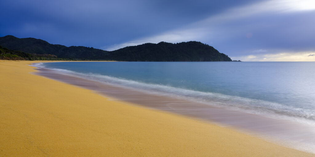 New Zealand, Nelson & Marlborough, Abel Tasman National Park. Dawn at the golden sand beach of Totaranui Bay which is bordered by native woodland and rocky outcrops.