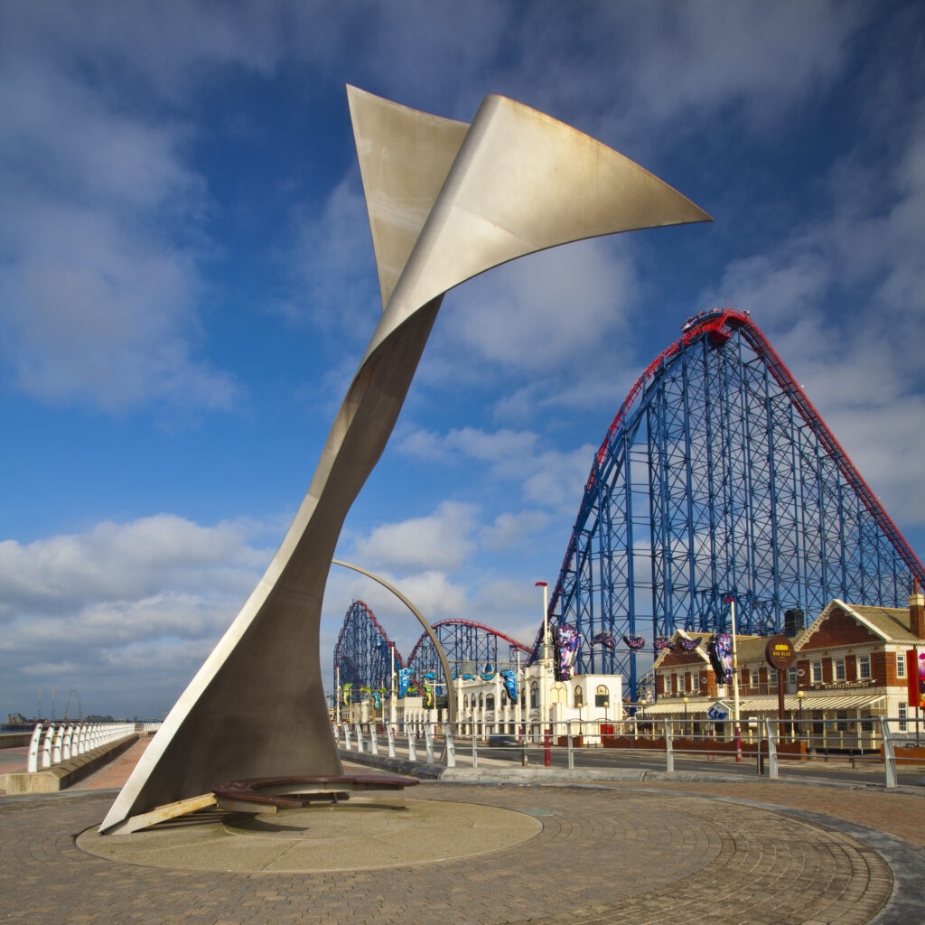 England, Lancashire, Blackpool. Whale tail sculpture on Blackpool's South Promenade with the Blackpool Pleasure Beach and the Big One (steel roller coaster) in the distance.