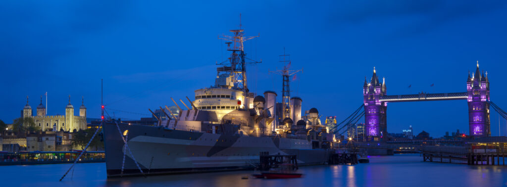 England, London, Southwark. HMS Belfast, originally a Royal Navy light cruiser, permanently moored in London on the River Thames with Tower Bidge and the Tower of London in the distance.
