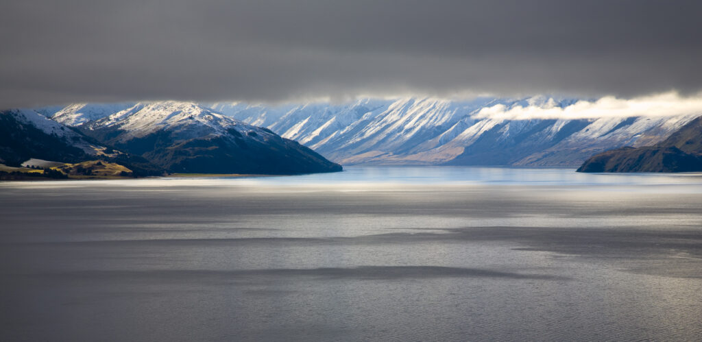 New Zealand, Otago, Lake Hawea. The Southern Alps revealed through a small gap between dense snow clouds and the surface of Lake Hawea.