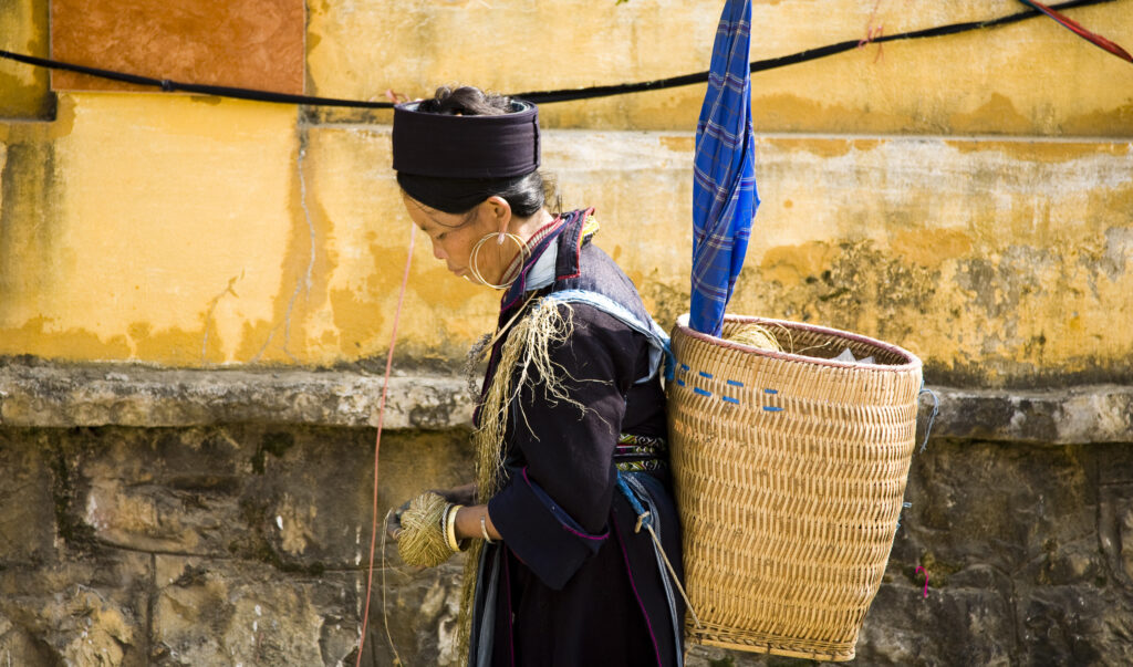 Vietnam, Northern Vietnam, Sapa. Local Sapa hill tribe woman with a basket on her back and twine wrapped around her hand, walking alongside a colourful wall Sapa.