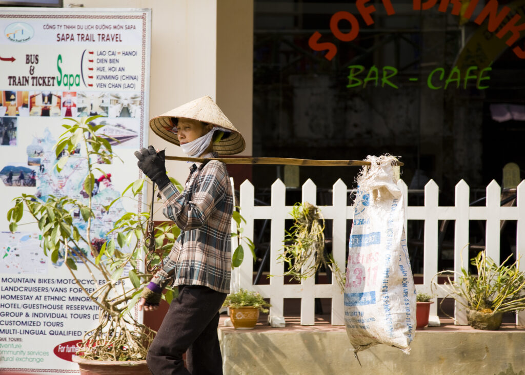 Vietnam, Northern Vietnam, Sapa. Local vietnamese woman carrying a sack on a stick resting on her shoulder walking past a bar-cafe in Sapa.