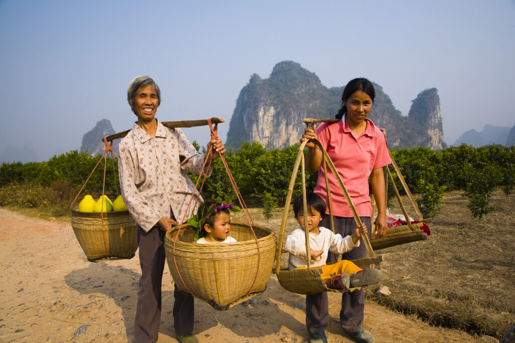 China, Guangxi Zhuang Autonomous Region, Yangshuo County. Two rural farm workers carry fruit and their children in hand woven baskets along a rural road running through an agriculture landscape dominated by the Karst peaks of Yangshuo County.