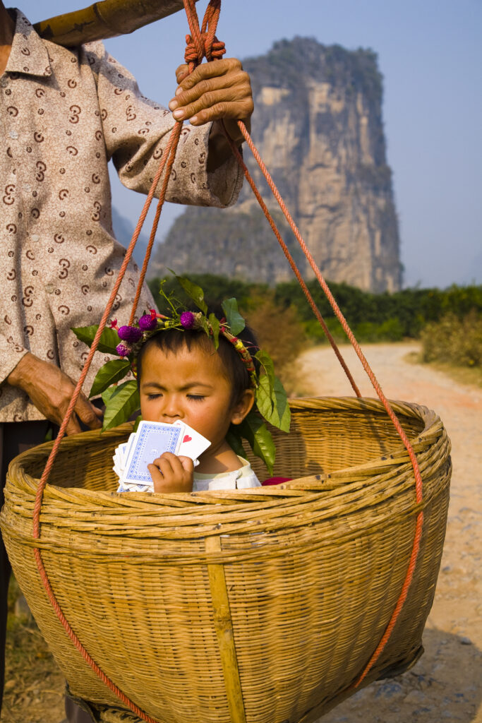 China, Guangxi Zhuang Autonomous Region, Yangshuo County. Young chinese child being carried in an hand woven basket along a rural road running through an agriculture landscape dominated by the Karst peaks of Yangshuo County.