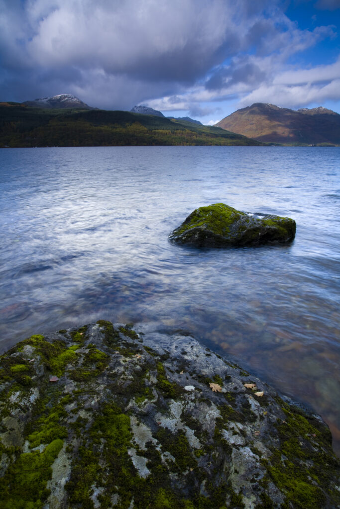 Scotland, Stirling, Loch Lomond and the Trossachs National Park. Moss covered boulders on Loch Lomond with mountains in the background.