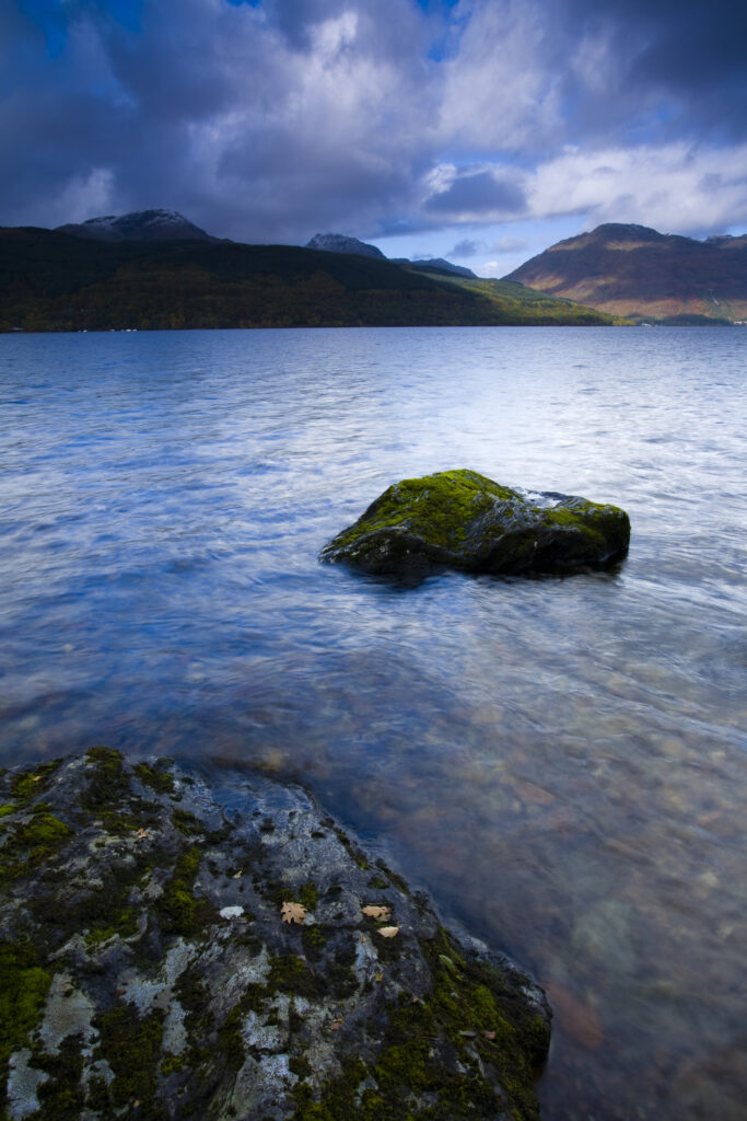 Scotland, Stirling, Loch Lomond and the Trossachs National Park. Moss covered boulders on Loch Lomond with mountains in the background.