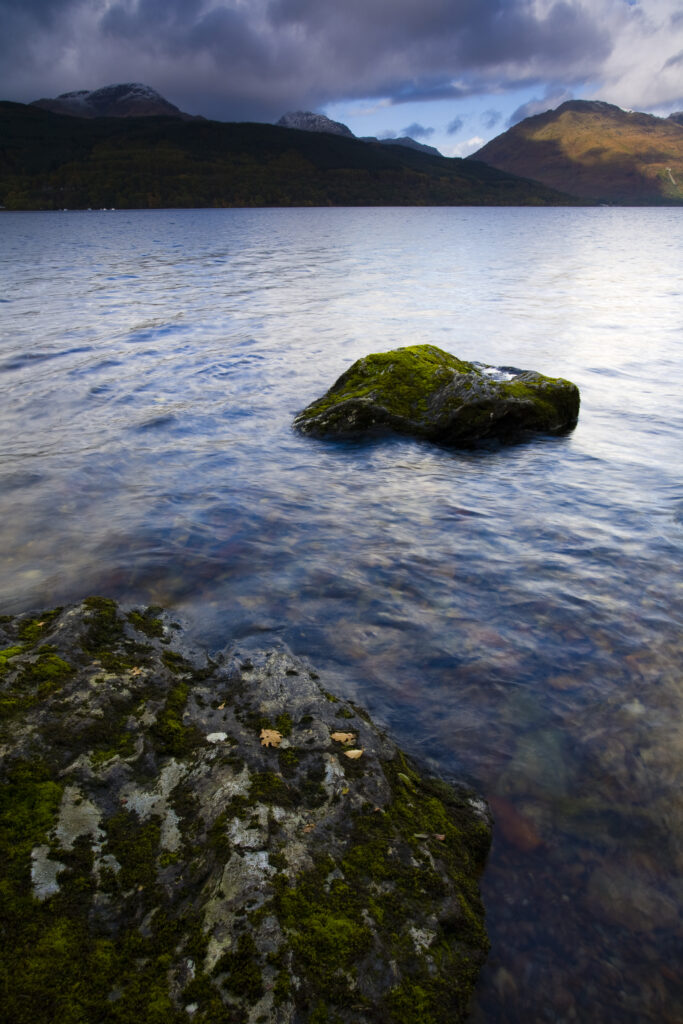 Scotland, Stirling, Loch Lomond and the Trossachs National Park. Moss covered boulders on Loch Lomond with mountains in the background.