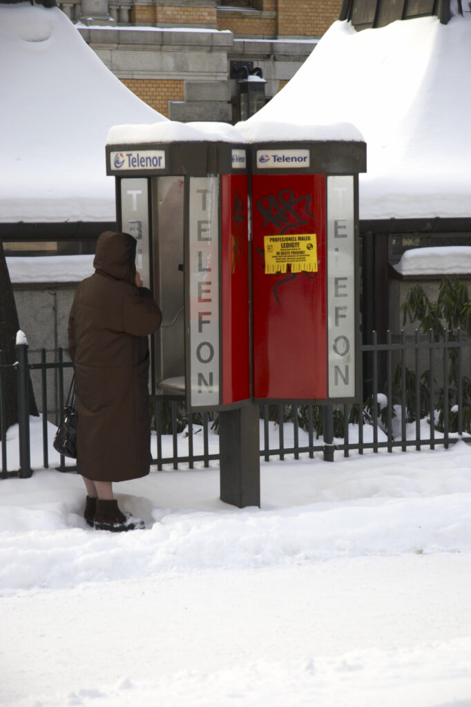 Norway, Oslo, Oslo City. An elderly lady uses a public phone booth in the snow covered streets of the city of oslo.