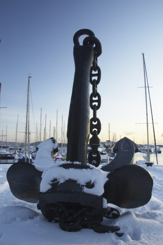 Norway, Oslo, Oslo City. Late afternoon winter light illuminates an anchor on the snow coverered Aker Quay and Oslofjorden Fjord