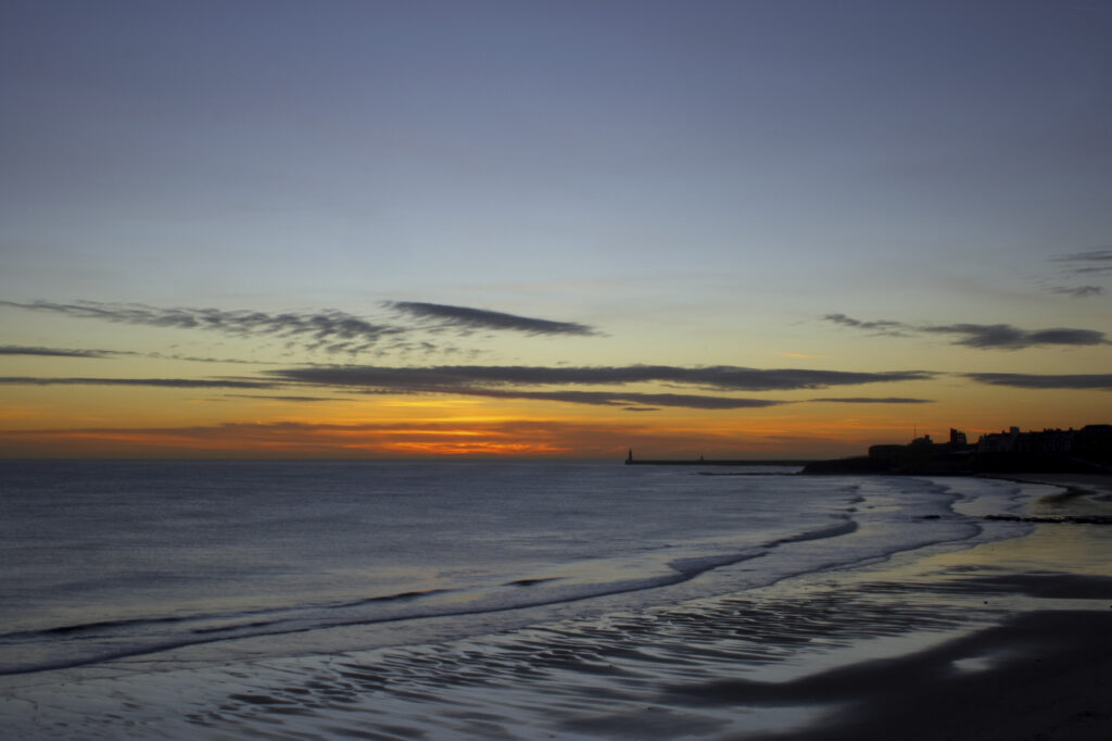 England, Tyne and Wear, Tynemouth. Sunrise looking across longsands towards the North Tyne Pier lighthouse and the Tynemouth priory.