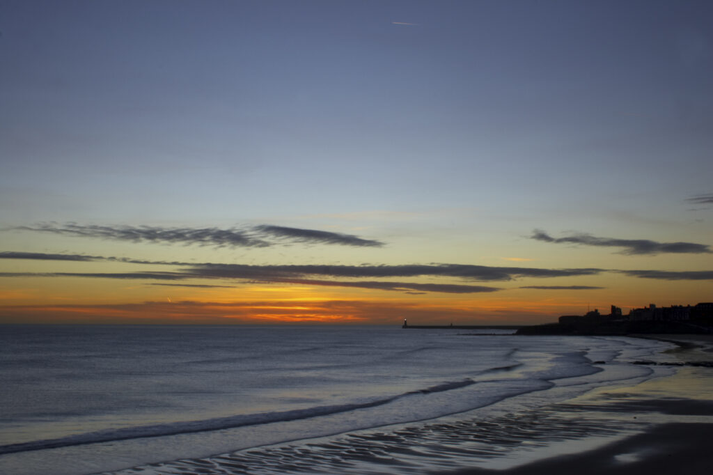 England, Tyne and Wear, Tynemouth. Sunrise looking across longsands towards the North Tyne Pier lighthouse and the Tynemouth priory.