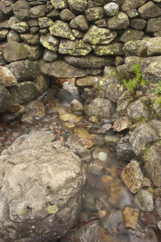 England, Cumbria, Lake District National Park. Stream running through a hole in drystone wall, in the Langdale Valley.