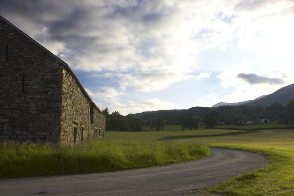 England, Cumbria, Lake District National Park. Soft evening light illuminates a small country lane and a stone farm building