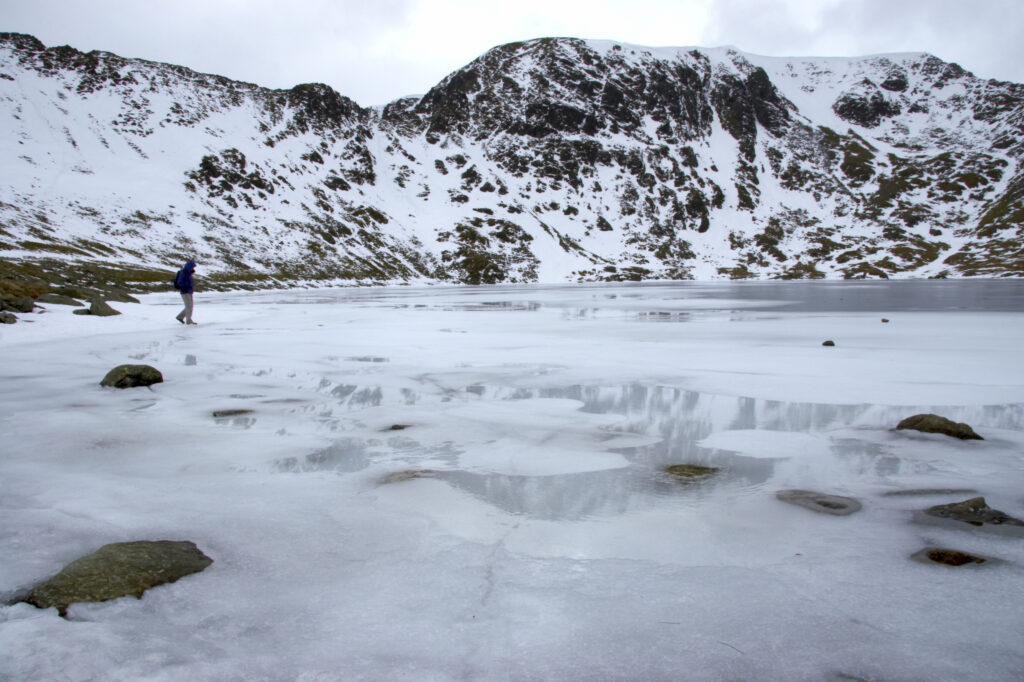 England, Cumbria, Lake District National Park. A hiker exploring the ice covered expanse of Red Tarn, beneath the snow capped peaks of Helvellyn.