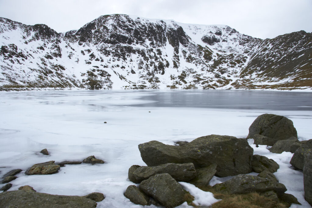 England, Cumbria, Lake District National Park. Rocks and the ice covered expanse of Red Tarn, beneath the snow capped peaks of Helvellyn.