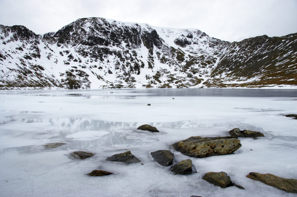 England, Cumbria, Lake District National Park. Rocks and the ice covered expanse of Red Tarn, beneath the snow capped peaks of Helvellyn.