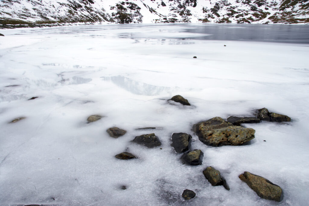 England, Cumbria, Lake District National Park. Rocks and the ice covered expanse of Red Tarn, beneath the snow capped peaks of Helvellyn.