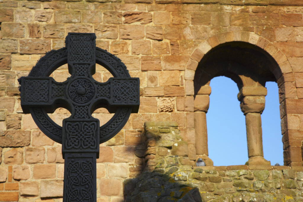 England, Northumberland, Lindisfarne / Holy Island. Celtic style grave stone against the ruins of the Lindisfarne Priory, located upon Holy Island along the Northumberland Heritage Coast.