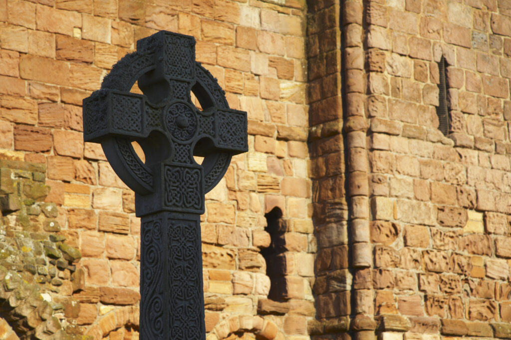 England, Northumberland, Lindisfarne / Holy Island. Celtic style grave stone against the ruins of the Lindisfarne Priory, located upon Holy Island along the Northumberland Heritage Coast.