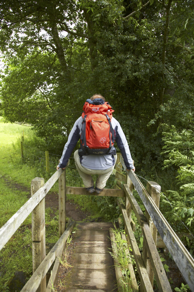 England, Cumbria, The Cumbria Way. A hiker jumps over a stile / bridge on the last day of the Cumbria Way track from Caldbeck to Carlisle.