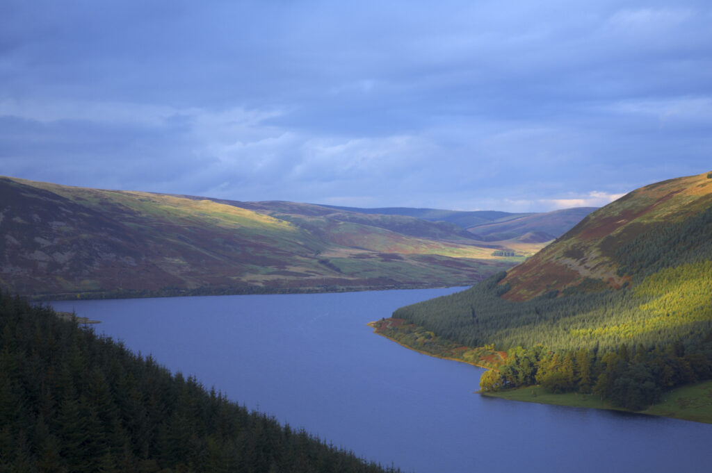 Scotland, Scottish Borders, St Marys Loch. Breaking light illuminates the autumn colours of the tree clad hills surrounding St Marys Loch.