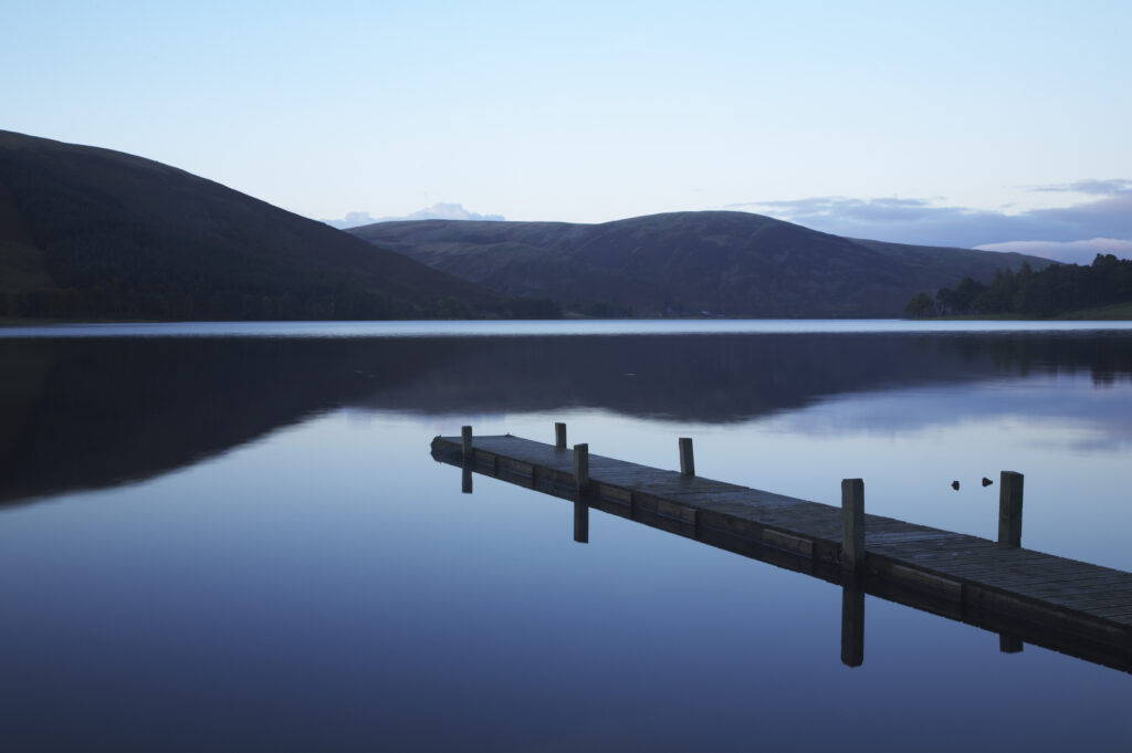 Scotland, Scottish Borders, St Marys Loch. St Marys Loch at dusk, with the surrounding hills and lakeside jetty reflected in the still waters of the loch.