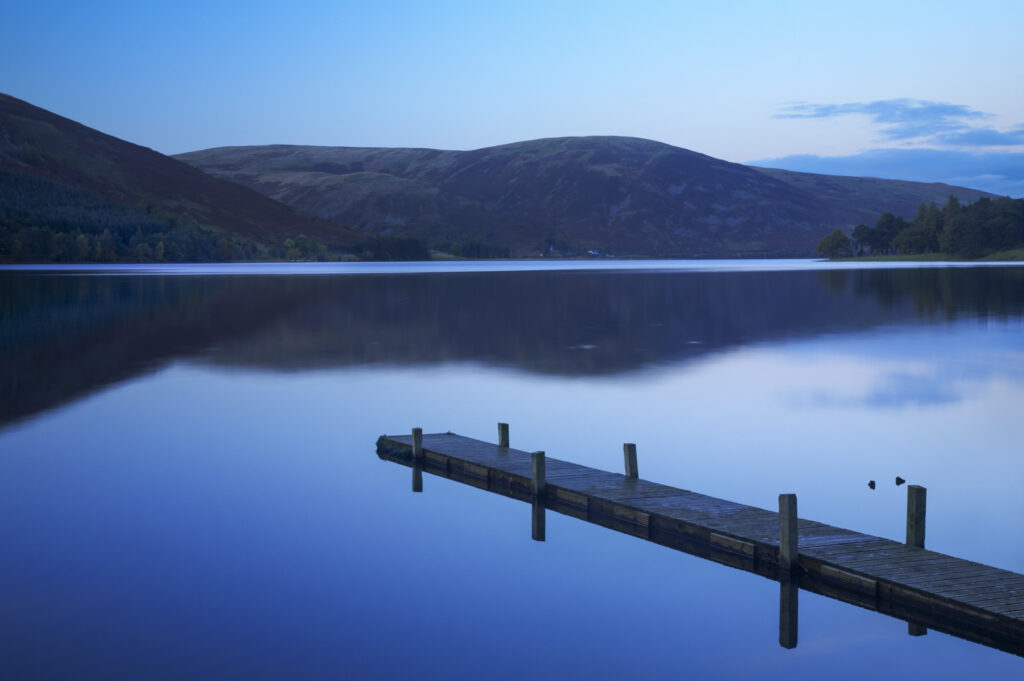 Scotland, Scottish Borders, St Marys Loch. St Marys Loch at dusk, with the surrounding hills and lakeside jetty reflected in the still waters of the loch.