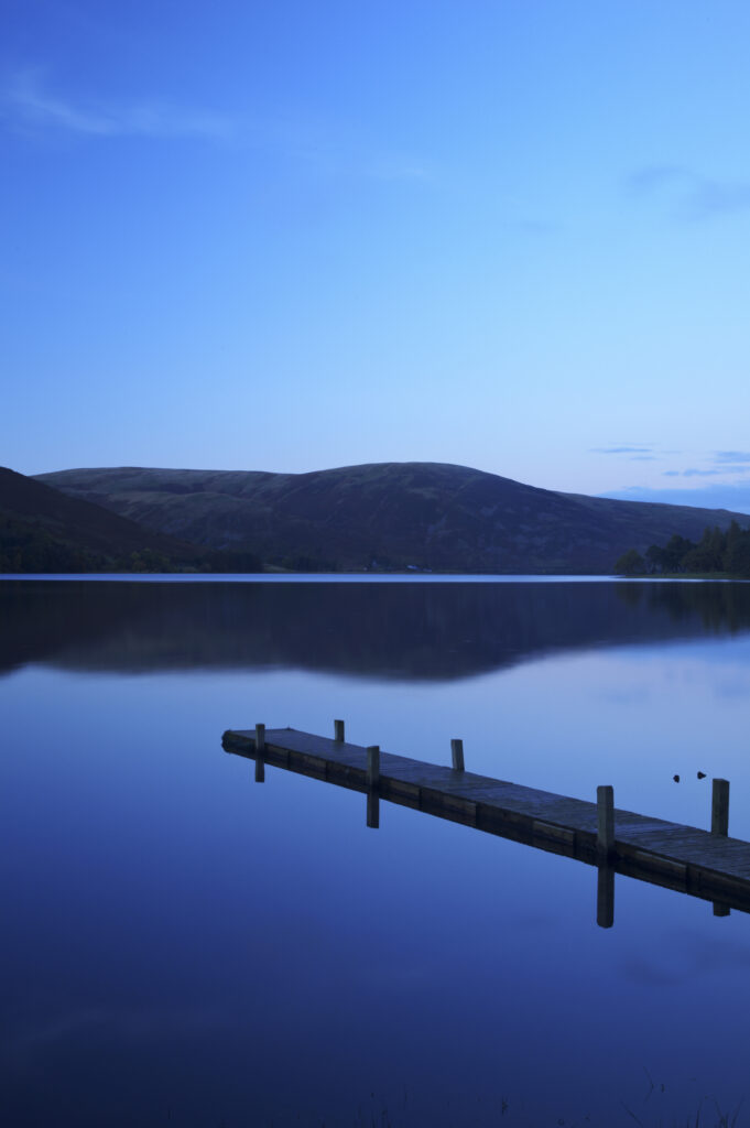 Scotland, Scottish Borders, St Marys Loch. St Marys Loch at dusk, with the surrounding hills and lakeside jetty reflected in the still waters of the loch.