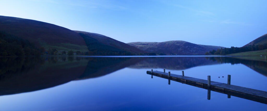 Scotland, Scottish Borders, St Marys Loch. St Marys Loch at dusk, with the surrounding hills and lakeside jetty reflected in the still waters of the loch.