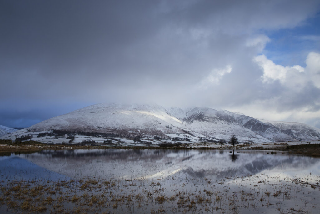 England, Cumbria, Lake District National Park. A winter view of Tewit Tarn (also spelt as Tewet Tarn) with a snow covered mountain (Blencathra) in the distance.