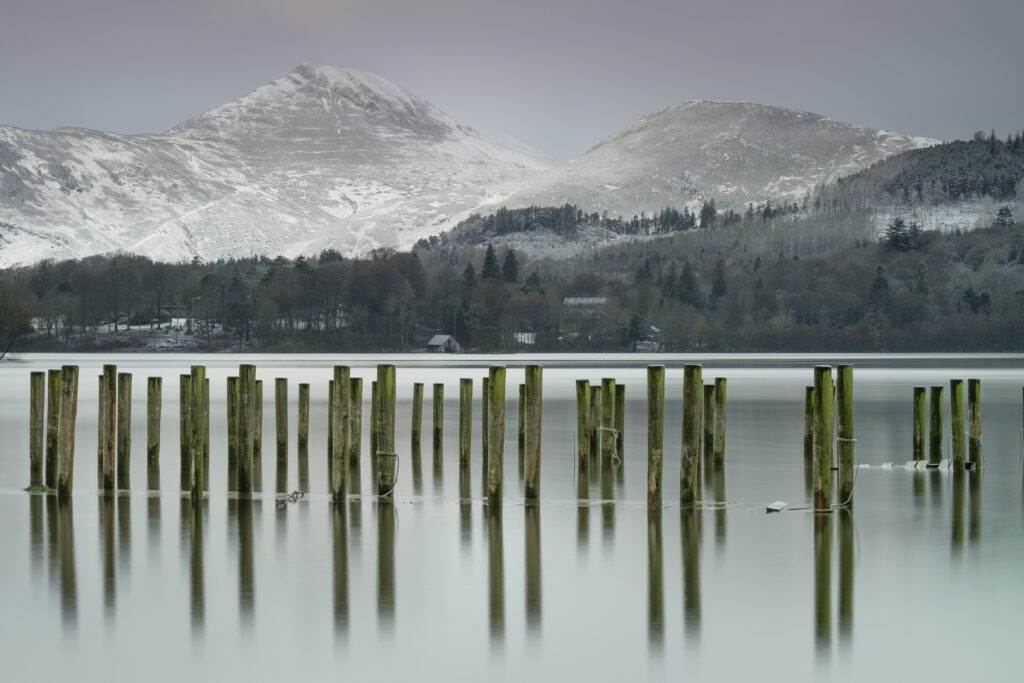 England, Cumbria, Lake District National Park. Mooring posts at the Keswick Jetty on Derwentwater with a snow covered mountain backdrop.