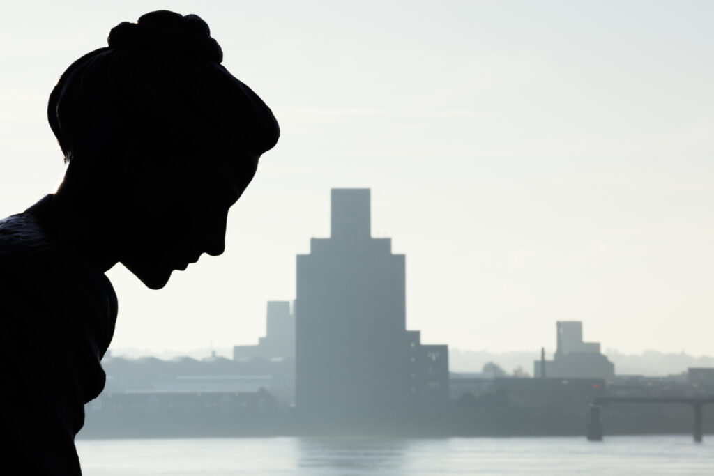 England, Merseyside, Liverpool. Statue located on the Liverpool Albert Docks known as 'The Crossing' with Birkenhead in the distance on the other side of the River Mersey.