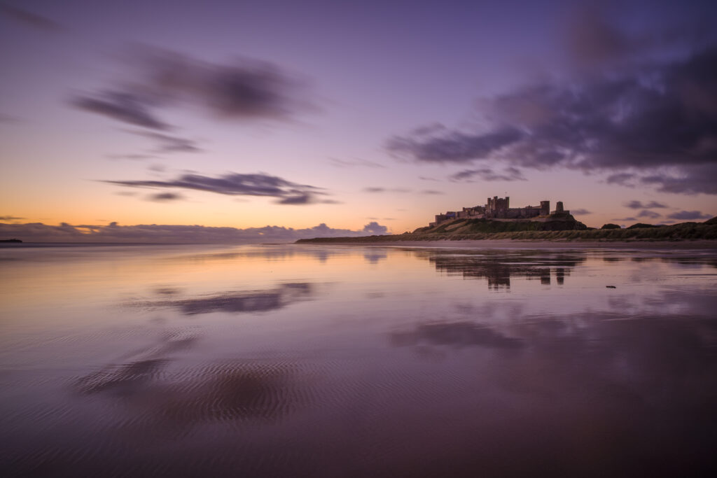 England, Northumberland, Bamburgh. The colours of dawn reflected on the wet, sandy beach at Bamburgh overlooked by Bamburgh Castle, located along the Northumberland Heritage Coast.