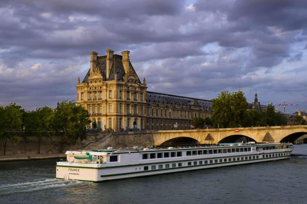 France, Paris, Louvre Palace. A tourist cruise ship on the River Seine heading towards the Pont Royal bridge overlooked by the Louvre Palace and museum.