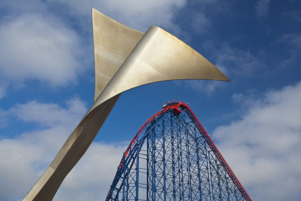 England, Lancashire, Blackpool. Whale tail sculpture on Blackpool's South Promenade with the Blackpool Pleasure Beach and the Big One (steel roller coaster) in the distance.