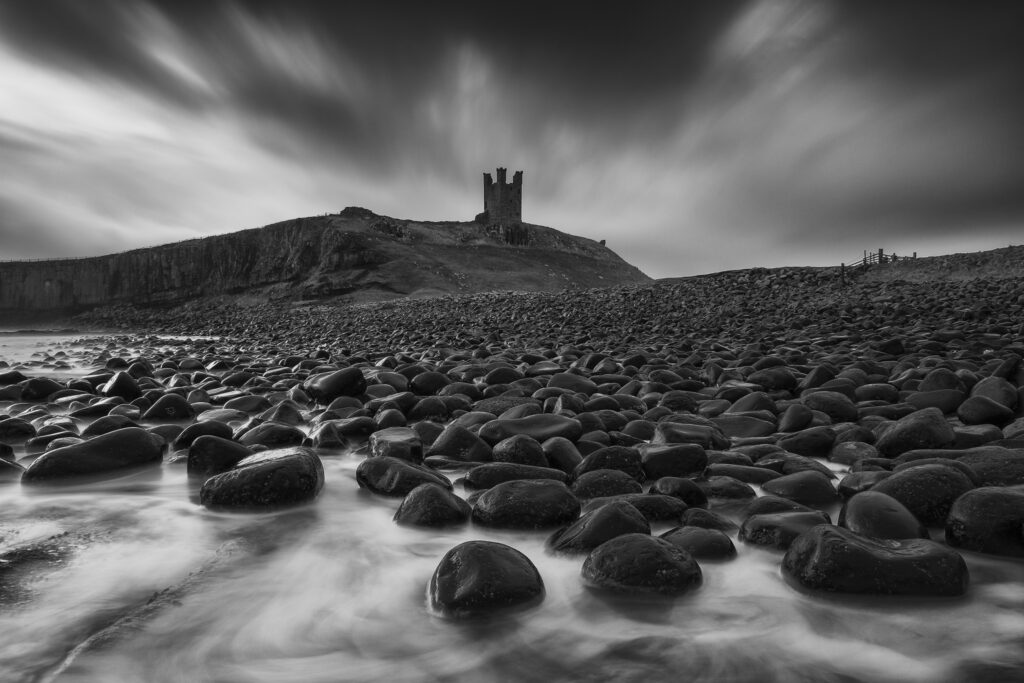 England, Northumberland, Embleton Bay. Waves crash againt the sea scultured rocks dominating the coastline of Embleton Bay, overlooked by the dramatic ruins of Dunstanburgh Castle.