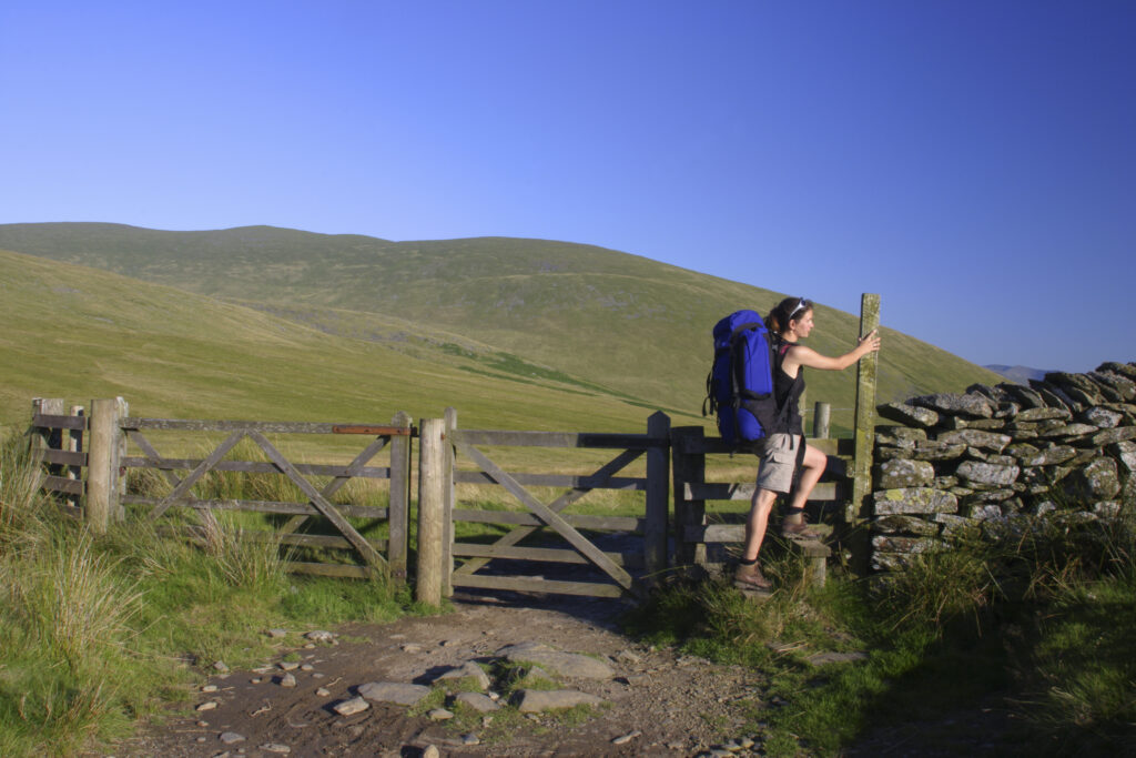 ENGLAND, Cumbria, Lake District National Park. A female hiker climbs a stile next to gate on track near Lonscale fell