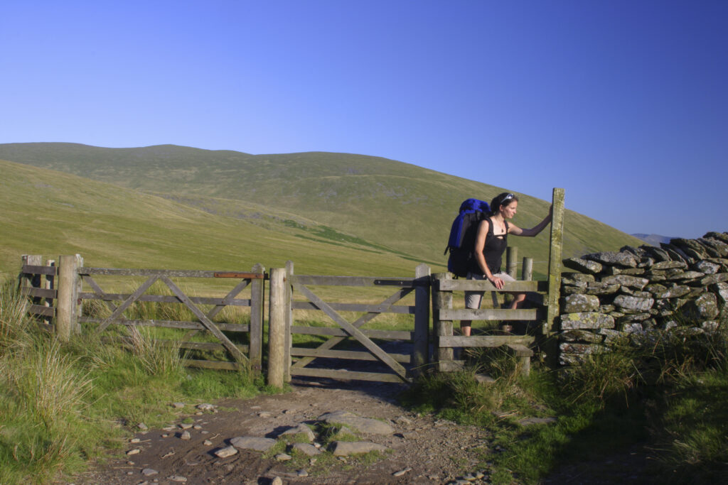 ENGLAND, Cumbria, Lake District National Park. A female hiker climbs a stile next to gate on track near Lonscale fell