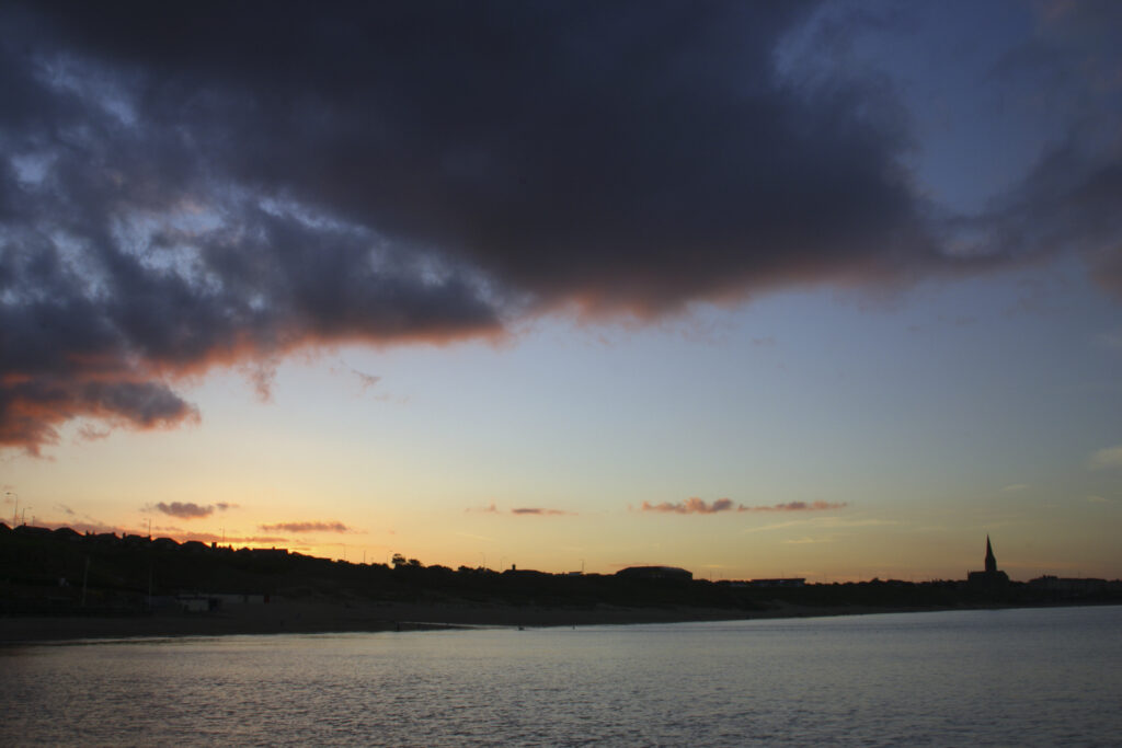 ENGLAND, Tyne & Wear, Tynemouth. The setting sun illuminates the clouds high above the long sands bay - looking towards Cullercoats