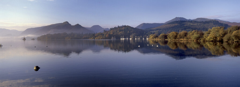ENGLAND, Cumbria, Lake District National Park. The surrounding autumn colours of woodland and mountain backdrop, reflected in the still waters of Derwentwater.