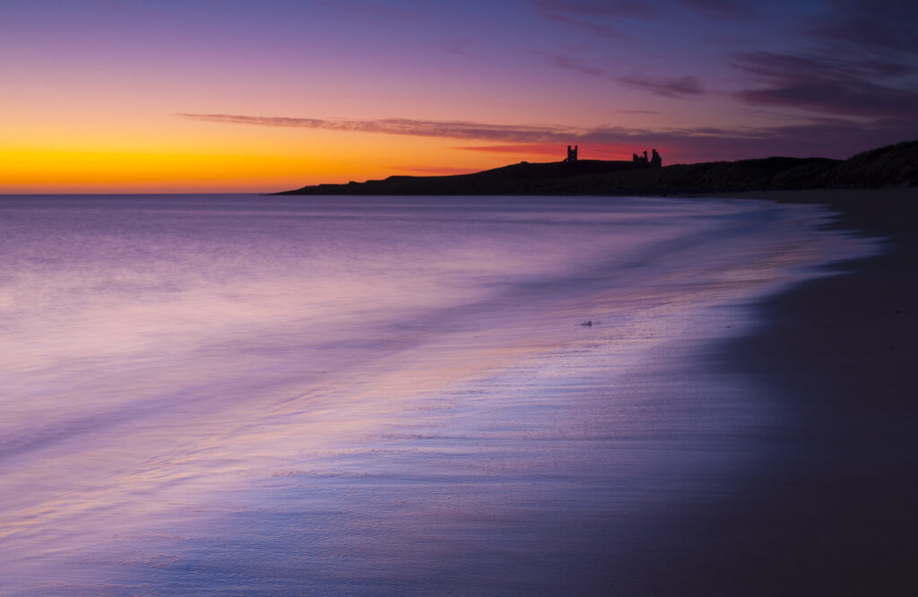 England, Northumberland, Embleton Bay. A colourful display of pre-dawn colours relected upon the wet sands of Embleton Bay, overlooked by the dramatic ruins of Dunstanburgh Castle.