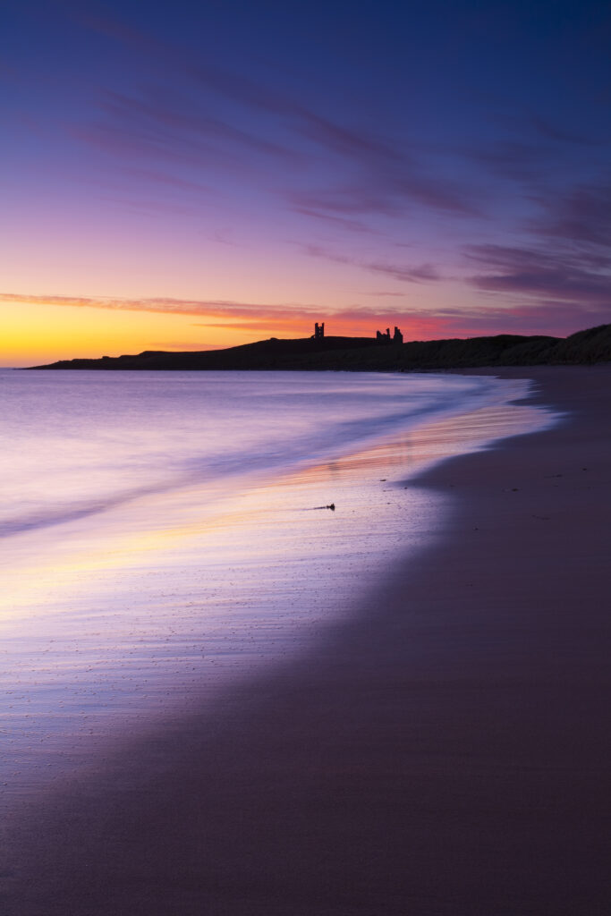 England, Northumberland, Embleton Bay. A colourful display of pre-dawn colours relected upon the wet sands of Embleton Bay, overlooked by the dramatic ruins of Dunstanburgh Castle.