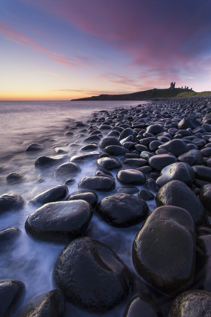 England, Northumberland, Embleton Bay. Waves crash againt the sea scultured rocks dominating the coastline of Embleton Bay, overlooked by the dramatic ruins of Dunstanburgh Castle.