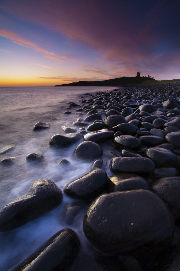 England, Northumberland, Embleton Bay. Waves crash againt the sea scultured rocks dominating the coastline of Embleton Bay, overlooked by the dramatic ruins of Dunstanburgh Castle.
