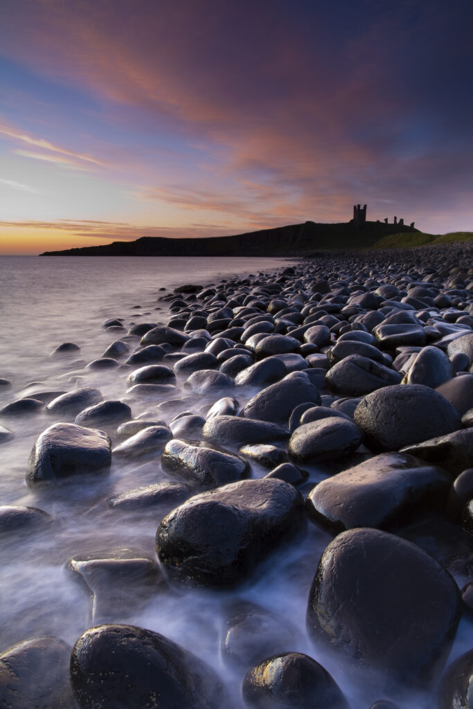 England, Northumberland, Embleton Bay. Waves crash againt the sea scultured rocks dominating the coastline of Embleton Bay, overlooked by the dramatic ruins of Dunstanburgh Castle.