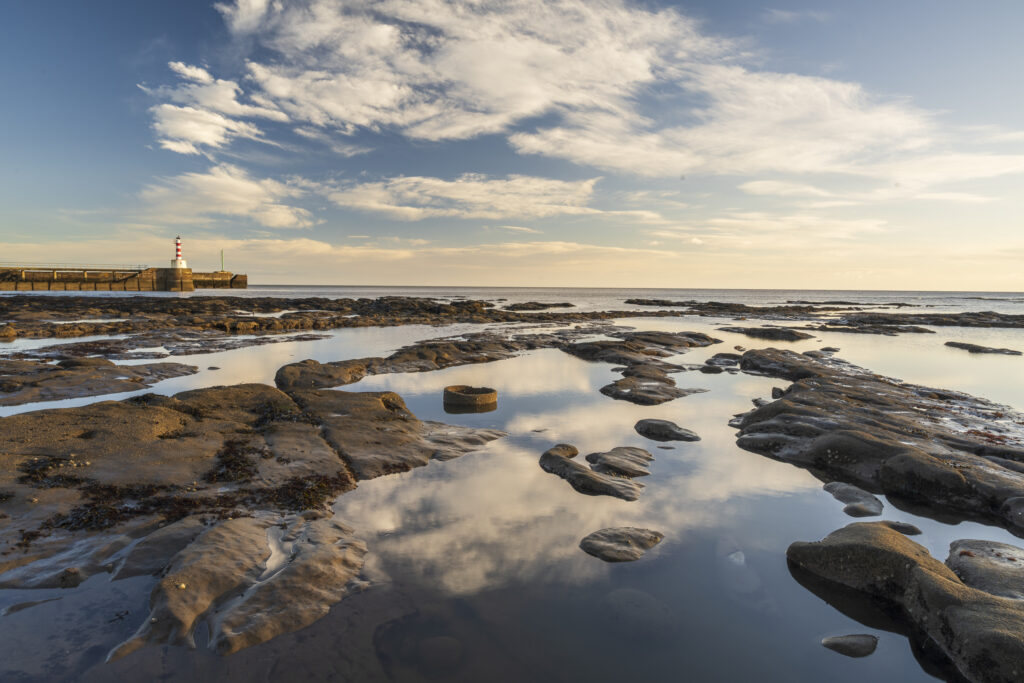 England, Northumberland, Amble. Early morning light illuminates the rocky shoreline near the Amble Harbour Pier.