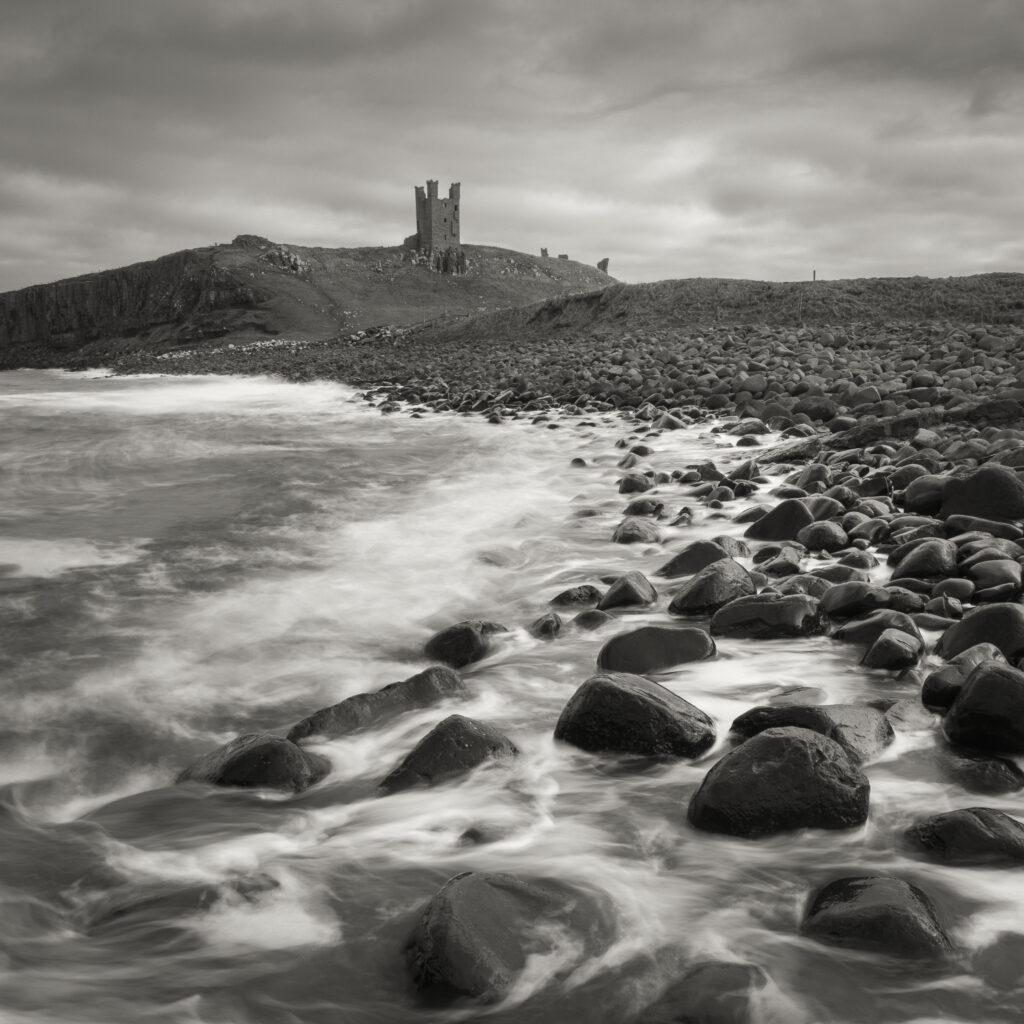 England, Northumberland, Embleton Bay. Waves crash againt the sea scultured rocks dominating the coastline of Embleton Bay, overlooked by the dramatic ruins of Dunstanburgh Castle.