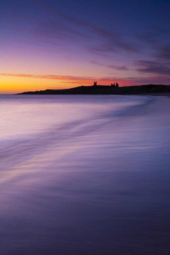 England, Northumberland, Embleton Bay. A colourful display of pre-dawn colours relected upon the wet sands of Embleton Bay, overlooked by the dramatic ruins of Dunstanburgh Castle.
